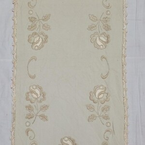 May include: A rectangular off-white table runner featuring light beige floral embroidery. The design includes five rose motifs arranged symmetrically, accented by curved scrollwork patterns. The edges are finished with a delicate crochet lace trim.