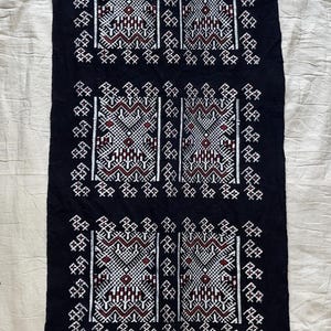 May include: A rectangular black textile runner featuring five repeating panels of intricate white and maroon geometric embroidery. The fabric has a fringed edge at both the top and bottom ends, showcasing a symmetrical pattern of lines and shapes.