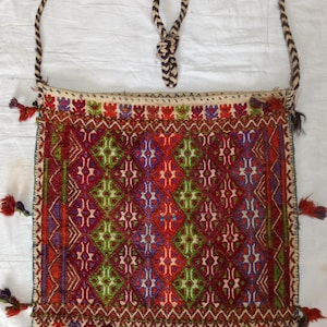 May include: A rectangular woven shoulder bag featuring a vibrant geometric pattern in red, green, purple, and white. The bag includes a braided strap, decorative tassels along the sides and bottom edge, and a complex diamond-shaped embroidery design.