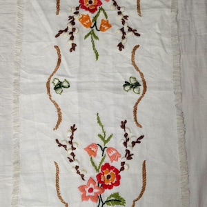 May include: A rectangular white linen table runner featuring fringed edges and a central embroidered design. The embroidery depicts two floral arrangements with red, orange, and pink flowers, green leaves, and brown stems, enclosed by a brown border.