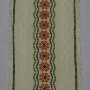 May include: A rectangular off-white table runner featuring a central vertical band of orange flowers with dark centres, bordered by green wavy lines. The fabric has a textured weave, side panels with subtle vertical patterns, and fringed ends.