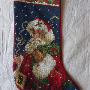 May include: A needlepoint Christmas stocking featuring a Santa Claus figure against a dark blue background with white snowflakes. The design includes a red border, a holly leaf trim at the top, and a red loop for hanging. Santa wears a red suit.