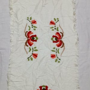 May include: A rectangular off-white table runner featuring a textured fabric surface with fringed edges on all sides. The runner is decorated with embroidered floral motifs in shades of red, orange, and green, arranged in a symmetrical pattern.