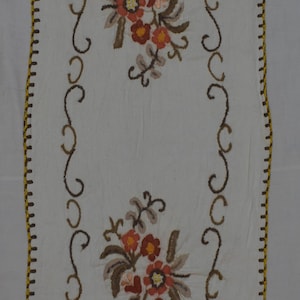 May include: A rectangular white fabric runner featuring two central floral embroidery motifs in shades of orange, brown, and yellow. The edges are finished with a decorative brown and yellow blanket stitch border and a brown embroidered scroll pattern.