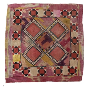 May include: A square textile piece featuring a central panel with four large diamond shapes in muted red and yellow tones. The border consists of a repeating geometric pattern of smaller squares in red, black, and cream, framed by a pink ikat fabric.