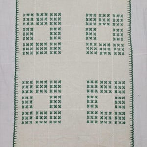 May include: A rectangular off-white table runner featuring a repeating pattern of eight green embroidered cross-stitch squares. The edges of the runner are finished with a decorative green blanket stitch border, creating a clean and structured look.