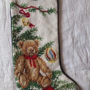 May include: A cream-coloured Christmas stocking featuring a cross-stitched design of a brown teddy bear with a red bow, green pine branches, a red bell, and colourful ornaments. The name Isaac is embroidered in dark green thread at the top.