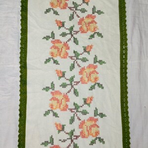 May include: A rectangular off-white table runner featuring a vertical cross-stitch pattern of orange and yellow roses with green leaves and stems. The runner is finished with a decorative dark green crocheted border along all four edges.