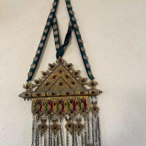 Puede incluir: Un colgante triangular de metal con grabados dorados, detalles en esmalte rojo y verde, e incrustaciones de piedras rojas. La pieza cuelga de una cinta de tela verde oscuro decorada con discos circulares plateados y pequeños cascabeles metálicos.