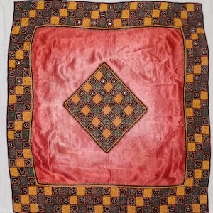 May include: A square textile piece featuring a solid coral-red center and a diamond-shaped central patch with a checkered pattern of yellow and dark green squares, each containing a small circular mirror. The border has a matching checkered design.