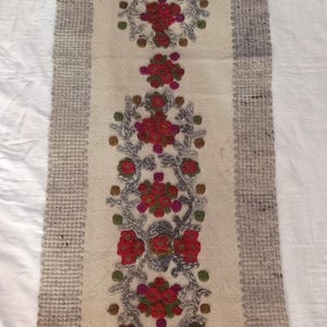 Puede incluir: Un camino de mesa largo y rectangular de color blanco roto con un patrón central vertical de motivos florales bordados en rojo y magenta, unidos por enredaderas grises y pequeños puntos verdes y marrones, con un borde gris y flecos.