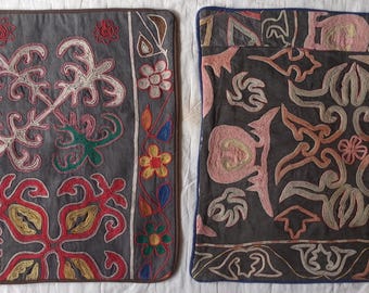 May include: Two square cushion covers featuring intricate embroidery on dark fabric. The left cover displays geometric and floral motifs in red, green, and yellow. The right cover features symmetrical, swirling patterns in shades of pink and beige.
