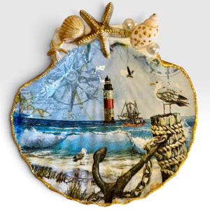 Op de afbeelding: Een decoratieve sint-jakobsschelp met een nautisch decoupage-ontwerp met een vuurtoren, een schip, een kompasroos en een meeuw. De rand van de schelp is goudkleurig geschilderd en aan de bovenkant versierd met een zeester, schelpjes en witte parels.