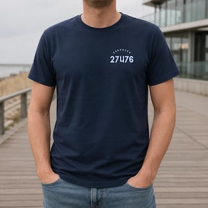 Könnte beinhalten: Ein dunkelblaues T-Shirt mit kurzen Ärmeln und Rundhalsausschnitt. Auf der linken Brustseite befindet sich ein weißer Schriftzug CUXHAVEN in einem Bogen über den großen weißen Ziffern 27476. Das T-Shirt besteht aus einfarbigem Stoff.