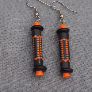 Peut inclure: Une paire de boucles d'oreilles avec des perles cylindriques noires et orange. Les perles ont un motif géométrique et sont attachées à des crochets argentés.