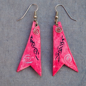 Peut inclure: Une paire de boucles d'oreilles triangulaires roses avec des motifs peints noirs et argentés. Les boucles d'oreilles ont une fermeture en crochet de fil d'argent.