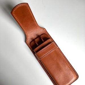 Op de afbeelding: Een bruinlederen pennenetui met een gestructureerde rechthoekige behuizing, een klepsluiting en zichtbare stiksels langs de randen. Het etui bevat interne verdelers om meerdere schrijfinstrumenten veilig in een rechtopstaande positie te houden.