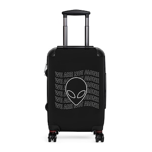 May include: Black rolling suitcase with a white alien head graphic and the text "WE ARE NOT ALONE". The luggage has a retractable handle and four wheels. The suitcase is designed for travel.