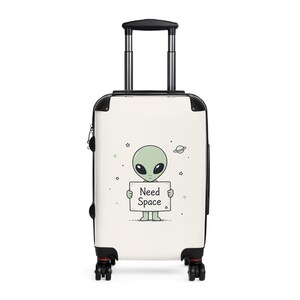 May include: A white rolling suitcase featuring a green alien holding a sign that reads "Need Space." The suitcase has a black handle and wheels, with a black and red trim. The design includes stars and a planet.