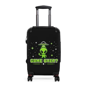 May include: Black rolling suitcase featuring a pixelated green alien in a spaceship, surrounded by stars. The phrase "GAME OVER?" is displayed in green pixelated text. The suitcase has a retractable handle and red accents.