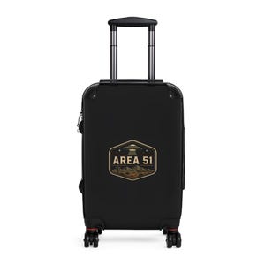 May include: Black rolling suitcase with a retractable handle. The suitcase features a graphic of a desert landscape with a UFO and the text "AREA 51" in a vintage style. The suitcase has red accents on the wheels.