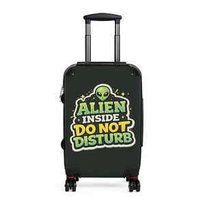 May include: A black rolling suitcase with a retractable handle. The front of the suitcase features a graphic of a green alien head and the text "ALIEN INSIDE DO NOT DISTURB" in yellow and green. The suitcase has black wheels and red accents.