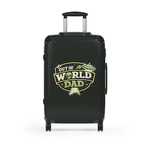 May include: A black rolling suitcase with a telescoping handle. The suitcase features a colorful graphic with the text "OUT OF WORLD DAD" and an alien and UFO design. The suitcase is designed for travel.