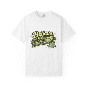 Op de afbeelding: Wit T-shirt met de tekst "Believe in Yourself (AND ALIENS)". Het ontwerp maakt gebruik van een retro lettertype in geel en groen, met een kleine UFO-illustratie. Het T-shirt heeft een klassieke ronde hals.