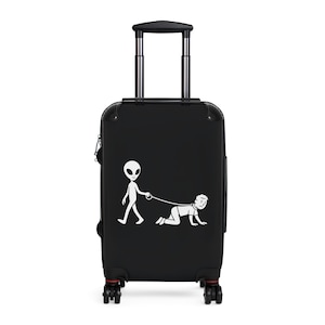 May include: Black rolling suitcase with a white graphic of an alien walking a person on a leash. The suitcase has a retractable handle and four wheels. The wheels and the bottom trim are red.
