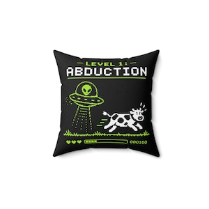 May include: Black square pillow with a pixelated design featuring a green alien in a UFO abducting a cow. The text reads "LEVEL 1: ABDUCTION" in white and green. A pixelated health bar and score are at the bottom.