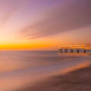 以下が含まれることがあります： 夕焼けの穏やかなビーチの風景。空は鮮やかな黄色から柔らかなピンクと紫に変化します。光の灯った桟橋が穏やかな水面に伸び、空の暖色を映し出しています。ビーチが前景にあります。