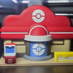 Könnte beinhalten: Ein 3D-gedrucktes Modell eines Pokémon-Center-Gebäudes mit rotem Dach und weißem Pokéball-Emblem, einer blauen Tür mit weißem Pokéball-Design, beigen Wänden, einem kleinen roten Kiosk mit blauem Bildschirm und einer gelben rechteckigen Kiste.