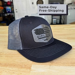 May include: A black baseball cap with a grey mesh back and a flat brim. The front features a grey hexagonal patch with a distressed American flag graphic, the text GIRL DAD, and the year EST. 2026 engraved in black on the center of the patch.