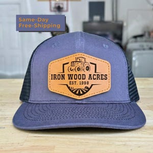 May include: A slate blue baseball cap with a black mesh back and a tan leather patch on the front. The patch features a black graphic of a tractor and the text IRON WOOD ACRES EST. 1998. The cap has a structured front panel and a curved brim.
