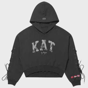 Op de afbeelding: Een zwarte cropped hoodie met de tekst GAP geborduurd op de capuchon en KAT by Magan in camouflageprint op de borst. De lange mouwen hebben zwarte vetersluitingen langs de buitenste naden en drie kleine nationale vlaggen op de boord.