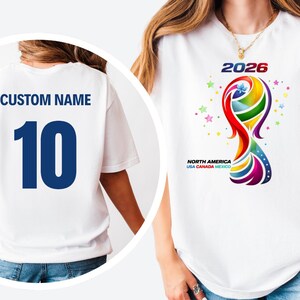 May include: A white t-shirt featuring a colorful 2026 graphic with the text North America USA Canada Mexico. The back of the shirt displays the text CUSTOM NAME and the number 10 in large dark blue font, shown in a circular inset overlay.