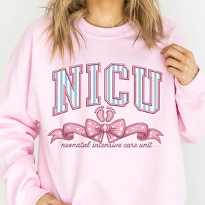 May include: A light pink crewneck sweatshirt featuring the text NICU in blue and white striped block letters. Below the text is a graphic of two small baby footprints and a pink ribbon with white hearts, with the text neonatal intensive care unit.