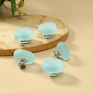May include: A set of four cabinet knobs featuring smooth, frosted light blue glass tops with an irregular, rounded shape. Each knob is attached to a polished silver-tone metal base, providing a clean and functional hardware component for furniture.