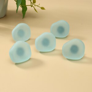 May include: Five smooth, rounded glass stones with a frosted, translucent light blue finish. Each stone features a darker, circular blue center, creating a layered appearance. The stones are arranged in a scattered formation on a flat surface.