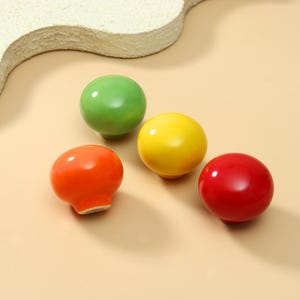 Op de afbeelding: Vier ronde, gladde meubelknoppen met een glanzende afwerking. De set bevat een groene, een gele, een oranje en een rode knop. Elke knop heeft een eenvoudige, paddenstoelachtige vorm met een afgeronde bovenkant en een kleine voet voor montage.