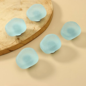 May include: A set of five cabinet knobs featuring smooth, rounded, frosted light blue glass tops with a matte finish. Each knob has a circular metal base, providing a secure attachment point for furniture drawers or cabinet doors.
