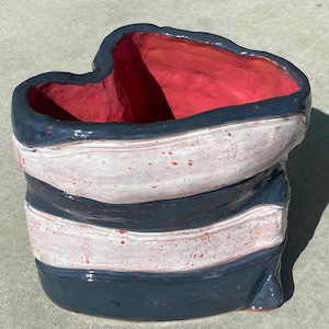 May include: A heart-shaped ceramic bowl featuring a glossy finish with alternating horizontal bands of dark charcoal gray and speckled white glaze. The interior of the bowl is finished in a vibrant, solid red glaze, creating a distinct contrast.