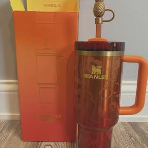 May include: A metallic orange Stanley Quencher H2.0 tumbler with a floral pattern, a gold-tone Stanley logo, and a gold pineapple straw topper. The tumbler includes a matching orange handle and lid, presented next to its orange and yellow packaging box.