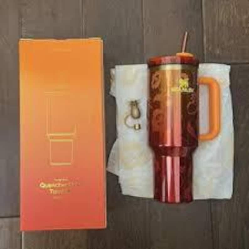 May include: A tall, metallic red Stanley Quencher H2.0 FlowState tumbler with an orange handle, a straw, and a gold logo. Beside it sits a matching orange box with the text Quencher H2.0 FlowState and a small gold-toned metal charm accessory.