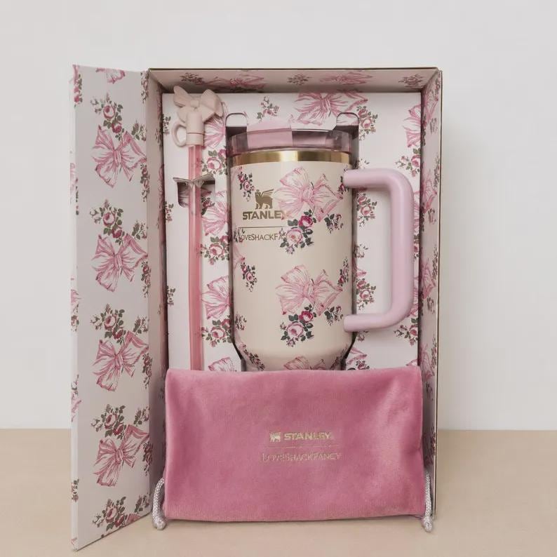 May include: A cream-colored Stanley tumbler with a pink floral and bow pattern, featuring a matching pink straw with a bow topper and a pink velvet pouch, all presented in a decorative box with a matching floral and bow print throughout.
