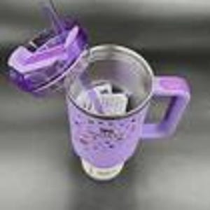 K&ouml;nnte beinhalten: Ein hellvioletter Isolierbecher mit Henkel und passendem violetten Deckel inklusive Strohhalm. Die Au&szlig;enseite des Bechers ist mit kleinen schwarzen grafischen Symbolen wie Sternen, Herzen und verschiedenen Formen verziert und hat ein metallisches Inneres.