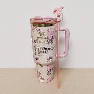 May include: A cream-colored Stanley Flowstate Quencher H2.0 tumbler with a 40 oz capacity, featuring a pink floral and ribbon pattern. It includes a matching pink handle, a clear lid, and a long pink straw topped with a small pink bow accessory.