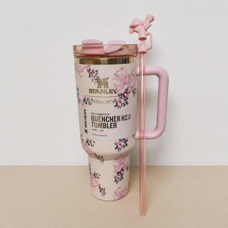 May include: A cream-colored Stanley Flowstate Quencher H2.0 tumbler with a 40 oz capacity, featuring a pink floral and ribbon pattern. It includes a matching pink handle, a clear lid, and a long pink straw topped with a small pink bow accessory.