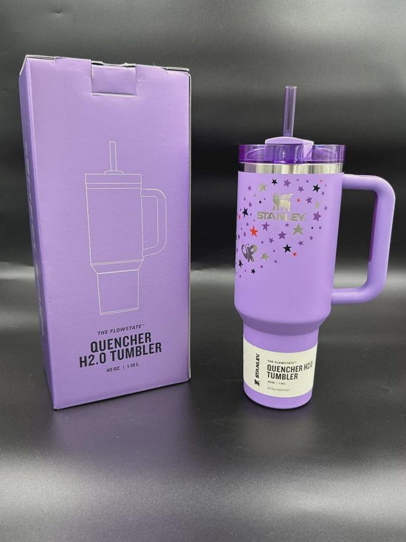 K&ouml;nnte beinhalten: Ein lavendelfarbener 1,18 L Stanley Quencher H2.0 Becher mit passendem Griff, Strohhalm und Deckel. Der Becher ist mit einem bunten Sternenmuster und dem Stanley-Logo versehen. Er steht neben der passenden violetten Verpackung The Flowstate Quencher H2.0.