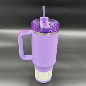K&ouml;nnte beinhalten: Ein hellvioletter Edelstahlbecher mit passendem Henkel, einem transparenten violetten Kunststoffdeckel und einem durchsichtigen Strohhalm. Der Becher hat ein wei&szlig;es rechteckiges Etikett nahe der Basis mit aufgedrucktem Text und Symbolen.